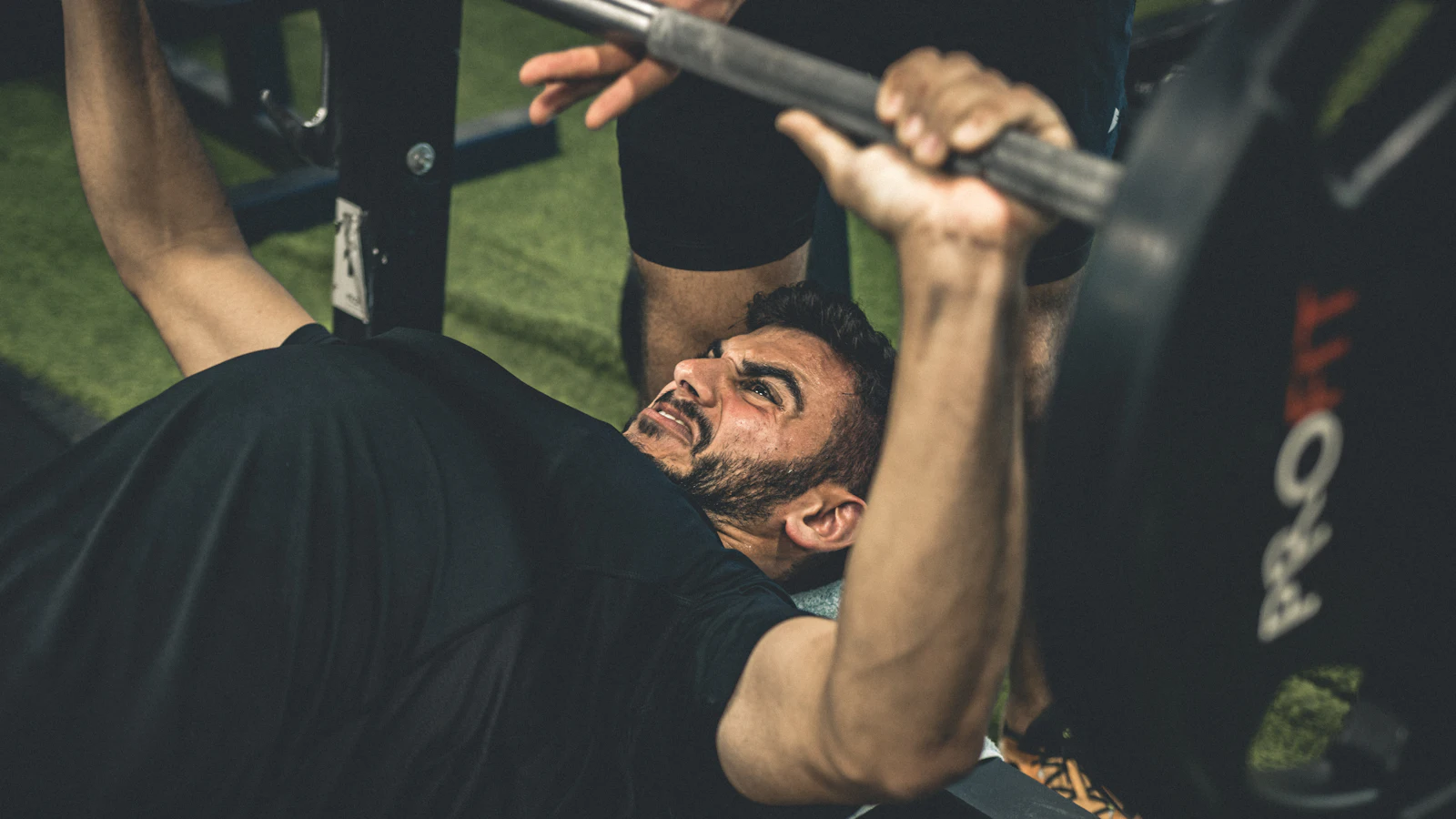 A man pressing a barbell on a bench while a training partner spots him