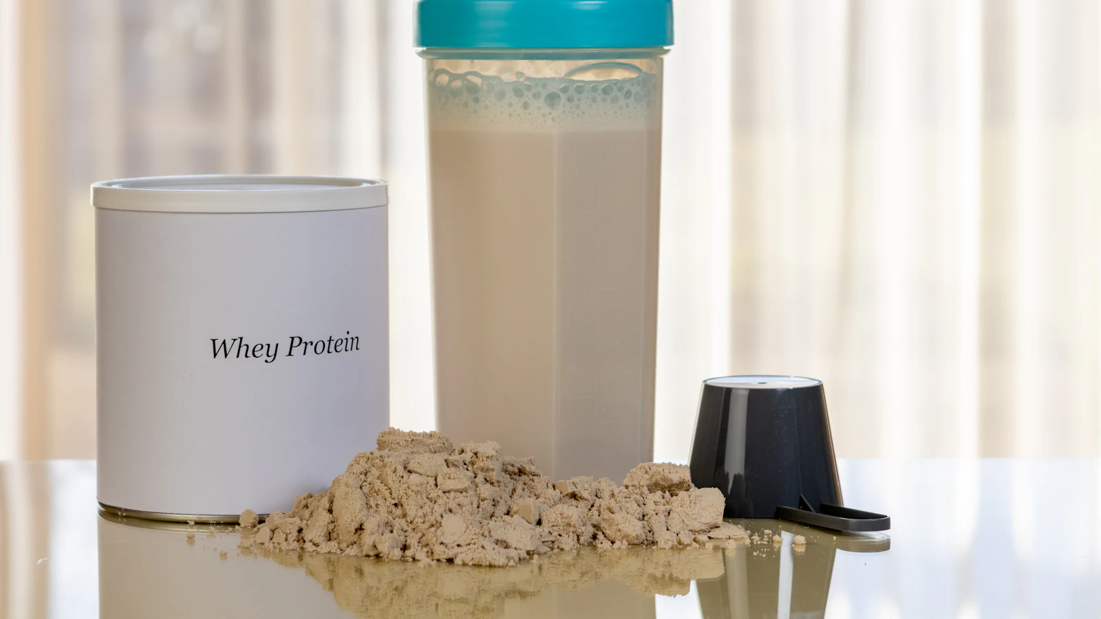 A tub labelled whey protein beside a shaker bottle of protein drink and a scoop